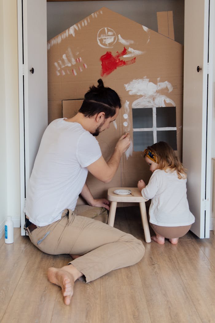 Father and daughter painting a cardboard house indoors, enjoying quality time together.